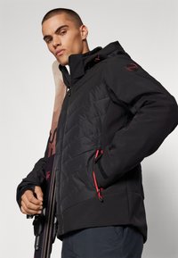 Schwarze Skijacke mit gestepptem Muster, verstellbarer Kapuze, roten Akzenten und Reißverschlüssen. Modell hält Skiausrüstung, neutraler Hintergrund.