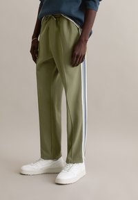 Olijfgroene sweatpants met witte en blauwe strepen aan de zijkant, een trekkoord in de tailleband en een zachte textuur, gecombineerd met witte sneakers.