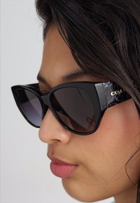 Schwarze Cat-Eye-Sonnenbrille mit Verlaufsgläsern, glänzenden Rahmen und Seitenverzierung. Logo sichtbar am Bügel mit texturiertem Finish.