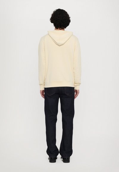 Cream-colored hoodie with a kangaroo pocket. Worn with dark denim jeans. The fabric appears soft with a relaxed fit and ribbed cuffs.