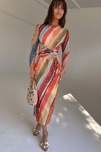 Színes, hosszú ujjú ruha, amely narancssárga, zöld, kék és sárga függőleges csíkokkal és pöttyökkel díszített; átlapolt kialakítású. Szőtt kézitáska és virágmintás magassarkúk.