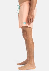 Shorts de bain pêche avec une taille élastique, cordon de serrage turquoise ajustable, fente sur les côtés et petit patch logo bleu en bas.