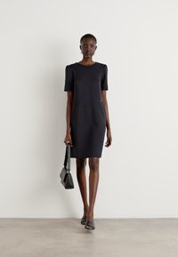 Robe noire à manches courtes avec une silhouette ajustée et des poches latérales. Présente un col rond et un tissu lisse. Mannequin portant un petit sac à main.