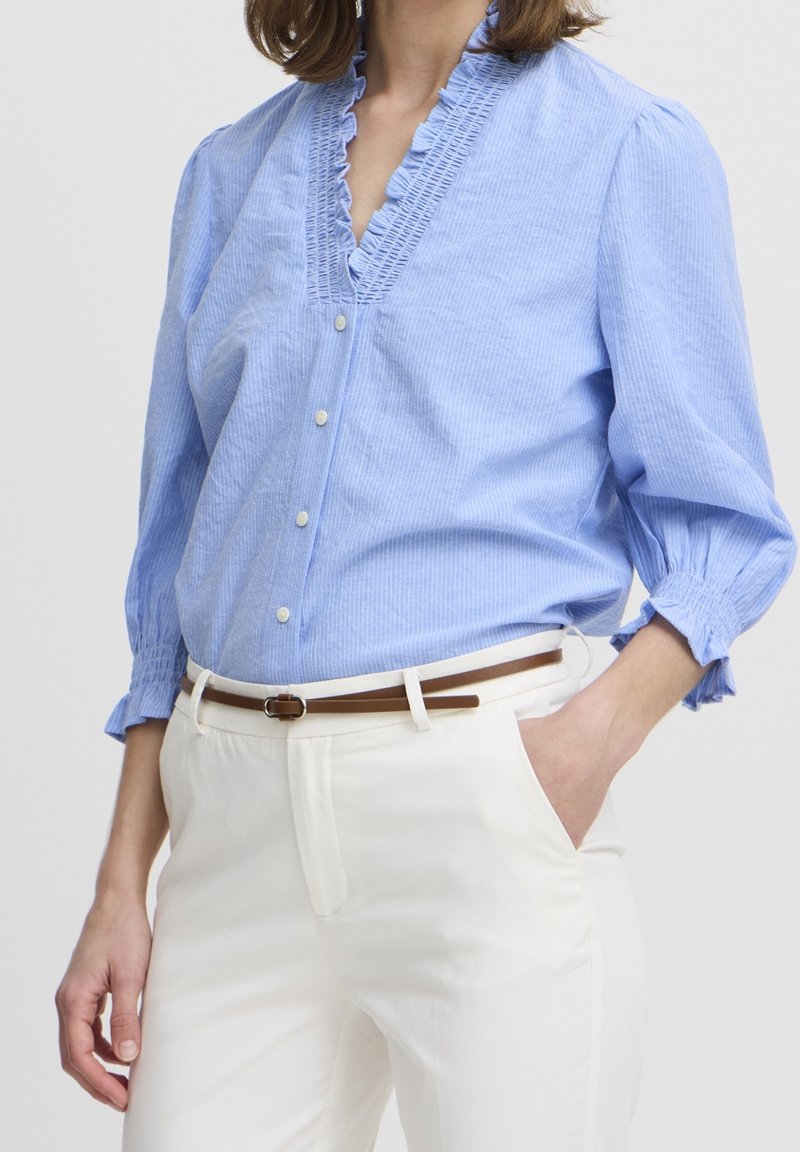 Femme portant un chemisier bleu clair à volants avec boutons et un pantalon blanc, ceinture marron, une main dans la poche, sur un fond uni.