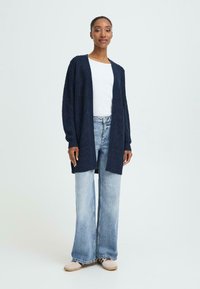 Mørkeblå strik cardigan med lange ærmer, åben front-design, parret med lyseblå klokkeformede jeans og beige fodtøj. Glat tekstur og afslappet pasform.