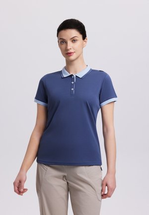 Kvinde med mørkt hår iført en blå polo med lyseblå krave og ærmekanter, iført beige bukser, stående mod en ensfarvet baggrund.