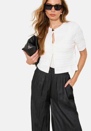 Vrouw die zwarte cat-eye zonnebril draagt, een wit topje met korte mouwen en knopen, zwarte wijde broekspijpen, en een zwarte clutch vasthoudt.