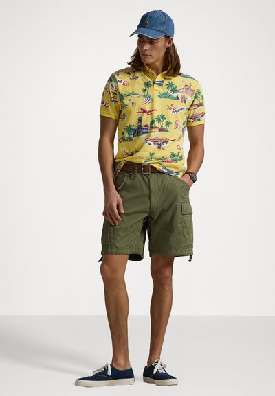 Camisa polo amarilla con estampados coloridos de aviación y tropicales, pantalones cortos de cargo verde oliva, zapatillas de lona azules y una gorra de denim.