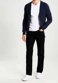 Mann trägt eine marineblaue Strickjacke über einem weißen V-Ausschnitt-Shirt, schwarze Hosen und weiße Sneakers, steht mit einer Hand in der Tasche.