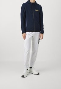Felpa zip da marina con tessuto testurizzato, tasche frontali e un'etichetta con il logo. Abbinata a pantaloni jogger bianchi con un design stampato e accentuazioni nere.