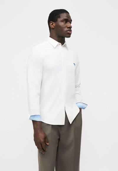 Άνδρας που στέκεται με τα χέρια στις τσέπες, φορώντας λευκό πουκάμισο με κουμπιά και μπλε ριγέ μανσέτες και καφέ παντελόνι, μπροστά σε απλό λευκό φόντο.