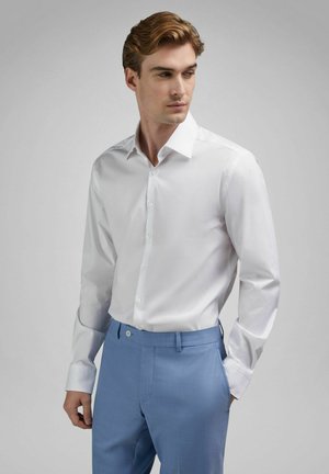 Man draagt een wit overhemd in een lichtblauwe, op maat gemaakte broek, staat met één hand in de zak tegen een grijze achtergrond.