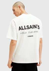 Fehér pamut póló, amelyen egy nagy fekete nyomat található a hátsó oldalon, "ALLSAINT'S" felirattal és további japán szöveggel, mellette pedig "LONDON" felirattal. Rövid ujjú.