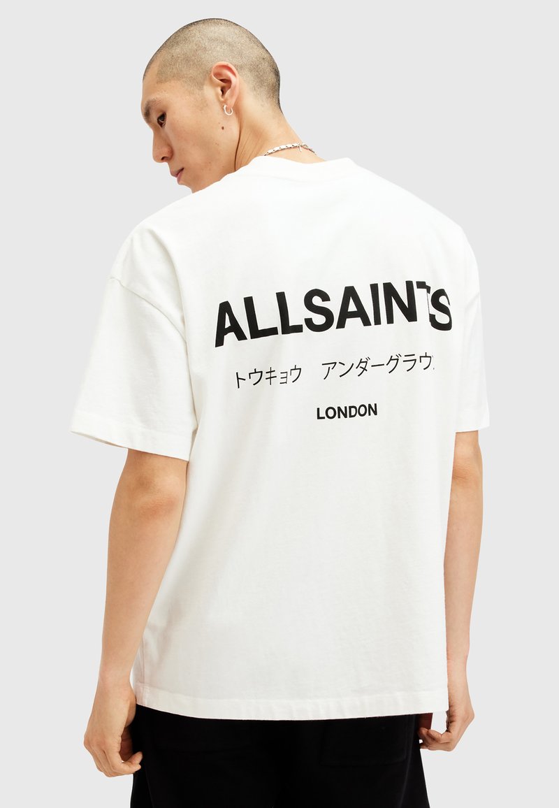 Fehér pamut póló, amelyen egy nagy fekete nyomat található a hátsó oldalon, "ALLSAINT'S" felirattal és további japán szöveggel, mellette pedig "LONDON" felirattal. Rövid ujjú.