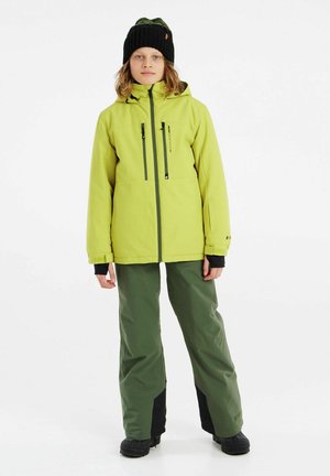 Veste imperméable verte éclatante avec poches zippées, associée à un pantalon vert ample avec des accents noirs et des bottes noires robustes.