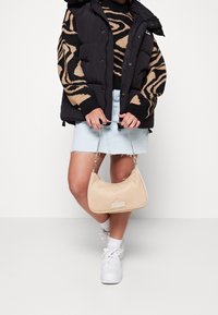 Beige handväska med en slät yta och silverkedjehandtag, hålls av en person som bär en svart pufferjacka och en mönstrad tröja.