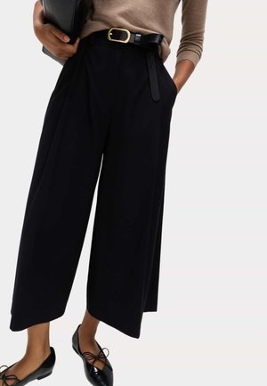 Person trägt schwarze Hosen mit weitem Bein, schwarzen Gürtel mit goldener Schnalle, beige langärmliges Oberteil und schwarze Ballerinas und hält eine schwarze Clutch-Tasche.