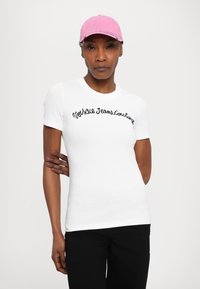 White fitted T-shirt with black embroidered text "Notable Jeans Couture." Paired with a pink cap. Simple design, short sleeves, smooth texture.