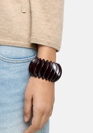 Nadgarstek z grubą, ciemnobrązową, zakrzywioną plastikową bransoletką, widoczny rękaw beżowego swetra i jasnoniebieskie dżinsy.