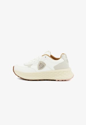 Sneaker bianca e beige con suola spessa e testurizzata, chiusura frontale con lacci, linguetta del tallone glitterata e toppa a forma di scudo decorata con paillettes sul lato.