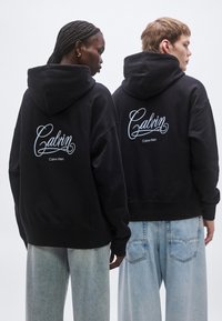 Čierne mikiny s kapucňou majú na chrbte veľké biele vyšívané logo "Calvin". Modelky majú na sebe voľné svetlomodré rifle z denimu.