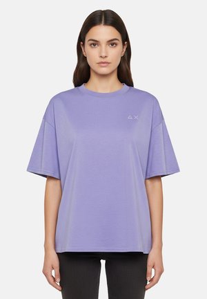 Donna con capelli lunghi e scuri che indossa una t-shirt lavanda a maniche corte e larghe e pantaloni neri, in piedi e rivolta verso l'obiettivo su uno sfondo bianco.
