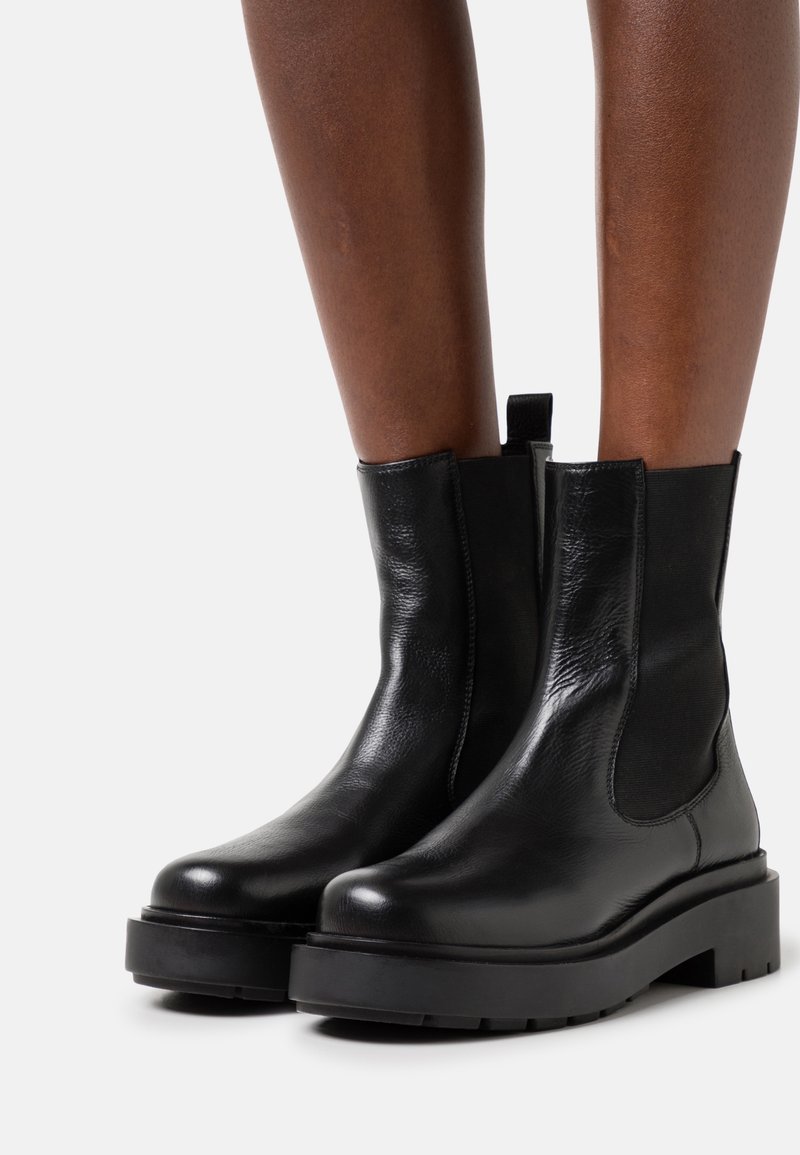 Bottines en cuir noir à semelles plateformes épaisses portées sur des jambes à peau foncée, avec des panneaux élastiques sur les côtés et une languette à l'arrière.