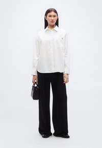 Λευκό πουκάμισο με κουμπιά και τσέπη, συνδυασμένο με άνετο μαύρο παντελόνι. Το μοντέλο κρατά μια μικρή μαύρη τσάντα, πάνω σε ουδέτερο φόντο.