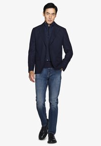 Marinblå tekstureret blazer over en marinblå lynlåsjakke, parret med lysblå denimjeans og sorte sko, hvilket skaber et lagdelt, moderne look.