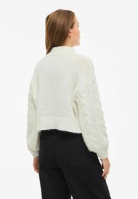 Bela pletena jopica z visokim rebrastim ovratnikom in skrajšanim krojem. Rokavi so rahlo napihnjeni, kar dodaja teksturo in dimenzijo.