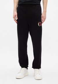 Μα�ύρες φόρμες από βαμβάκι, με ελαστική ζώνη μέσης και κόκκινο και λευκό λογότυπο στο αριστερό μηρό. Συνδυασμένες με άσπρα αθλητικά παπούτσια.