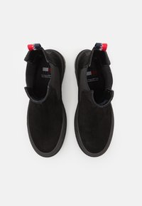 O pereche de botine negre din suede cu părți elastice și lupturi roșii, albe și albastre pe un fundal alb.