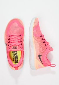 Paire de chaussures de course Nike roses vives avec un swoosh noir, des accents pêche et des semelles blanches sur un fond blanc.