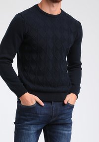 Granatowy sweter dzianinowy z wzorem diamentowym, okrągłym dekoltem oraz prążkowanymi mankietami i brzegiem. Noszony z dżinsami.