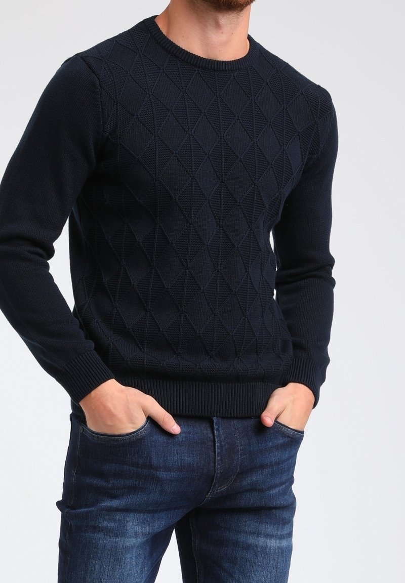 Granatowy sweter dzianinowy z wzorem diamentowym, okrągłym dekoltem oraz prążkowanymi mankietami i brzegiem. Noszony z dżinsami.