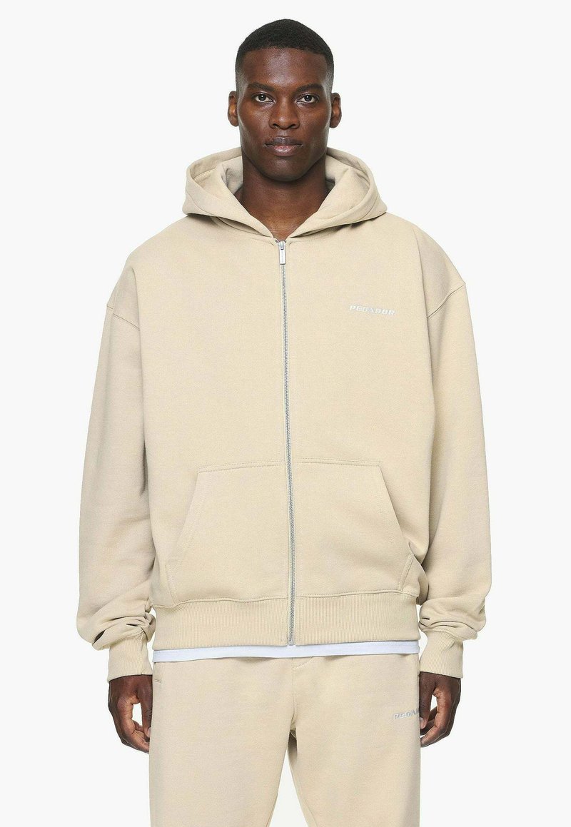 Beige zip-up hettegenser laget av myk stoff, med snøring i hetten, framlommer, og diskret merkevare på brystet.