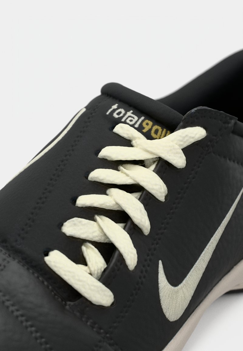 Zapatilla negra con cordones blancos, logo de swoosh blanco de Nike y texto "total 90" en la lengüeta, mostrada en primer plano sobre un fondo blanco.