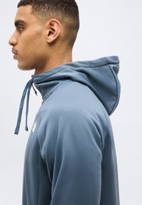 Νεαρός άνδρας σε προφίλ φορόντας ανοιχτό μπλε φούτερ με κουκούλα και κορδόνια, με φόντο απλό λευκό.