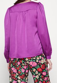 Blusa di raso viola con maniche a sbuffo e pieghe posteriori, abbinata a pantaloni neri floreali con motivi rosa, rossi e verdi.