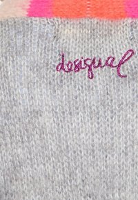 Tissu tricoté gris arborant le mot "desigual" brodé en violet. La partie supérieure présente un dégradé de rose vif à orange.