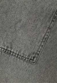 Tessuto denim grigio con una trama texturizzata, caratterizzato da cuciture visibili lungo i bordi e una tasca nell'angolo in alto a sinistra.