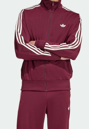 Osoba ubrana w bordowy dres Adidas z białymi paskami na rękawach i spodniach, stoi z założonymi rękami.