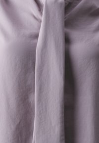 Lilac blouse with a tied neckline detail. Soft fabric with a smooth texture and draped fit, creating subtle folds in the material.