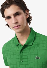 Polo verde con un motivo a griglia testurizzato, due bottoni al colletto e un piccolo logo di coccodrillo sul lato sinistro del petto.