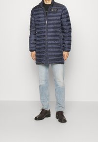 Marinblå puffjacka, quiltad design, hög krage, dragkedjestängning, broderad logotyp på bröstet. Matchad med ljusa jeans och mörka skor.