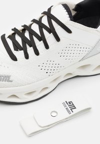 Scarpa sportiva bianca realizzata in mesh testurizzato con lacci neri. Presenta una suola spessa con intagli e un'etichetta bianca con marchio.