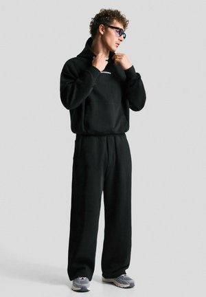 Man met krullend haar die een zwarte hoodie aanpast, met bijpassende zwarte sportbroek en grijze sneakers, staand tegen een effen lichte achtergrond.