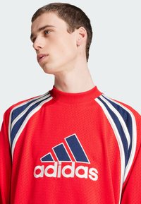 Haut de sport long rouge avec col ras du cou, texture en maille, rayures bleues et blanches sur les épaules, et un logo Adidas blanc bien en vue.