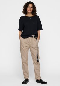 Vrouw met krullend haar, draagt een zwarte top met korte mouwen, beige broek met een zwarte riem en zwarte instappers, staat met haar handen in de zakken.