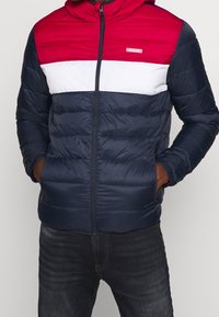 Zateplená bunda s kapucí v barevných blocích červené, bílé a námořnické modré. Vyrobena z lehkého materiálu, má přední zip a boční kapsy.